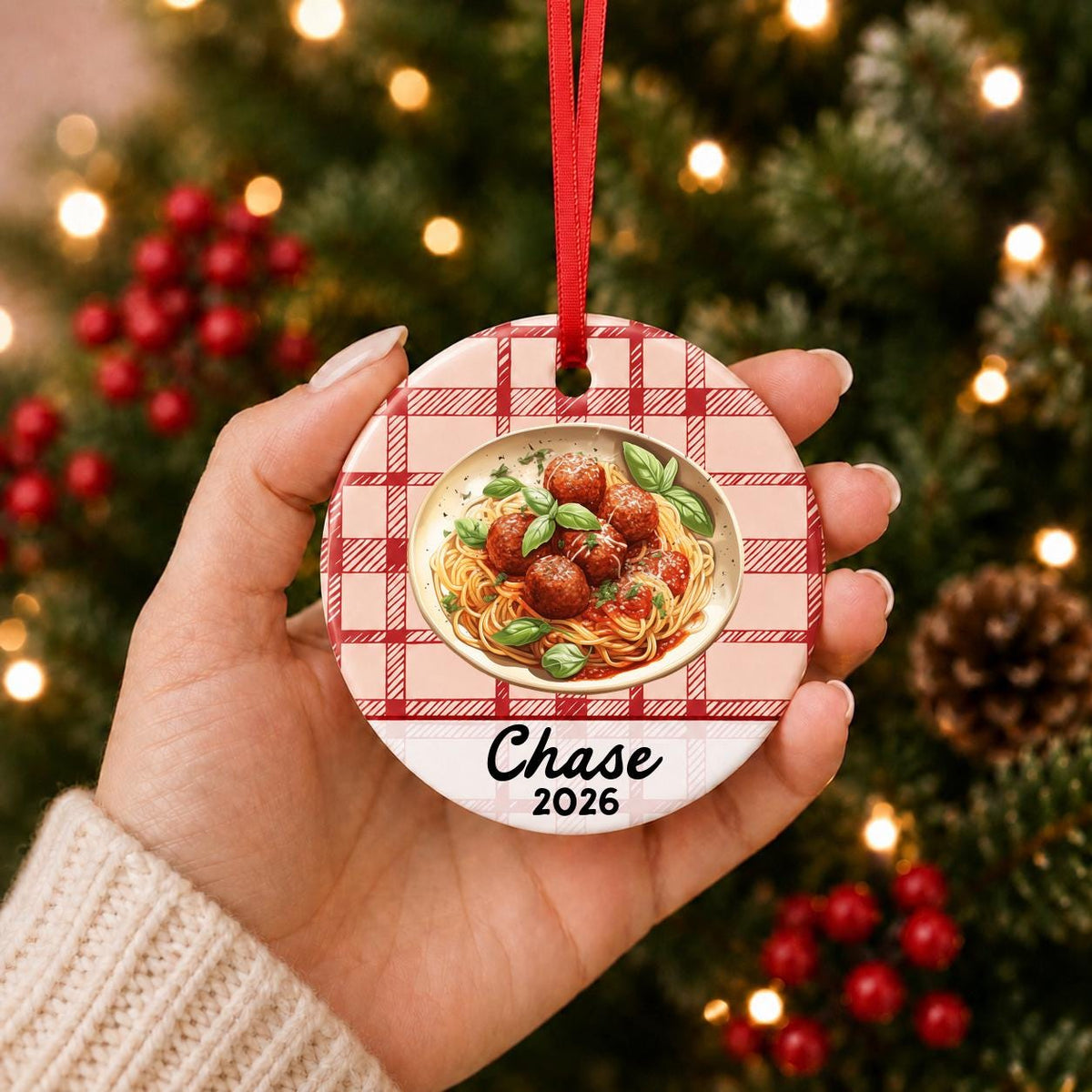 Hand holding the personalized spaghetti and meatballs ceramic ornament with name and year on red plaid in front of a Christmas tree