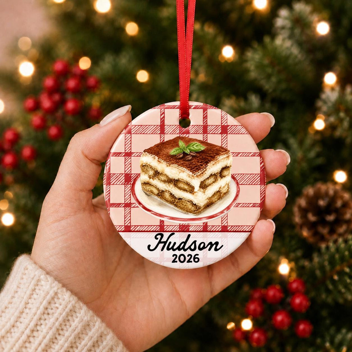 Hand holding the personalized tiramisu ceramic ornament with name and year on red plaid in front of a Christmas tree