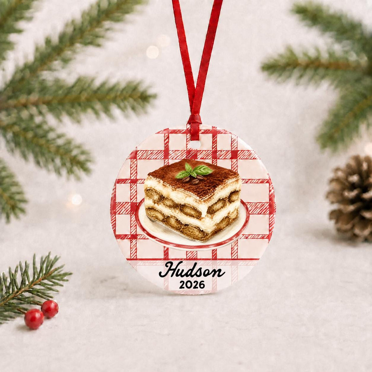 Personalized tiramisu ceramic ornament with name and year on red plaid with red ribbon on a white surface with pine branches and pinecones
