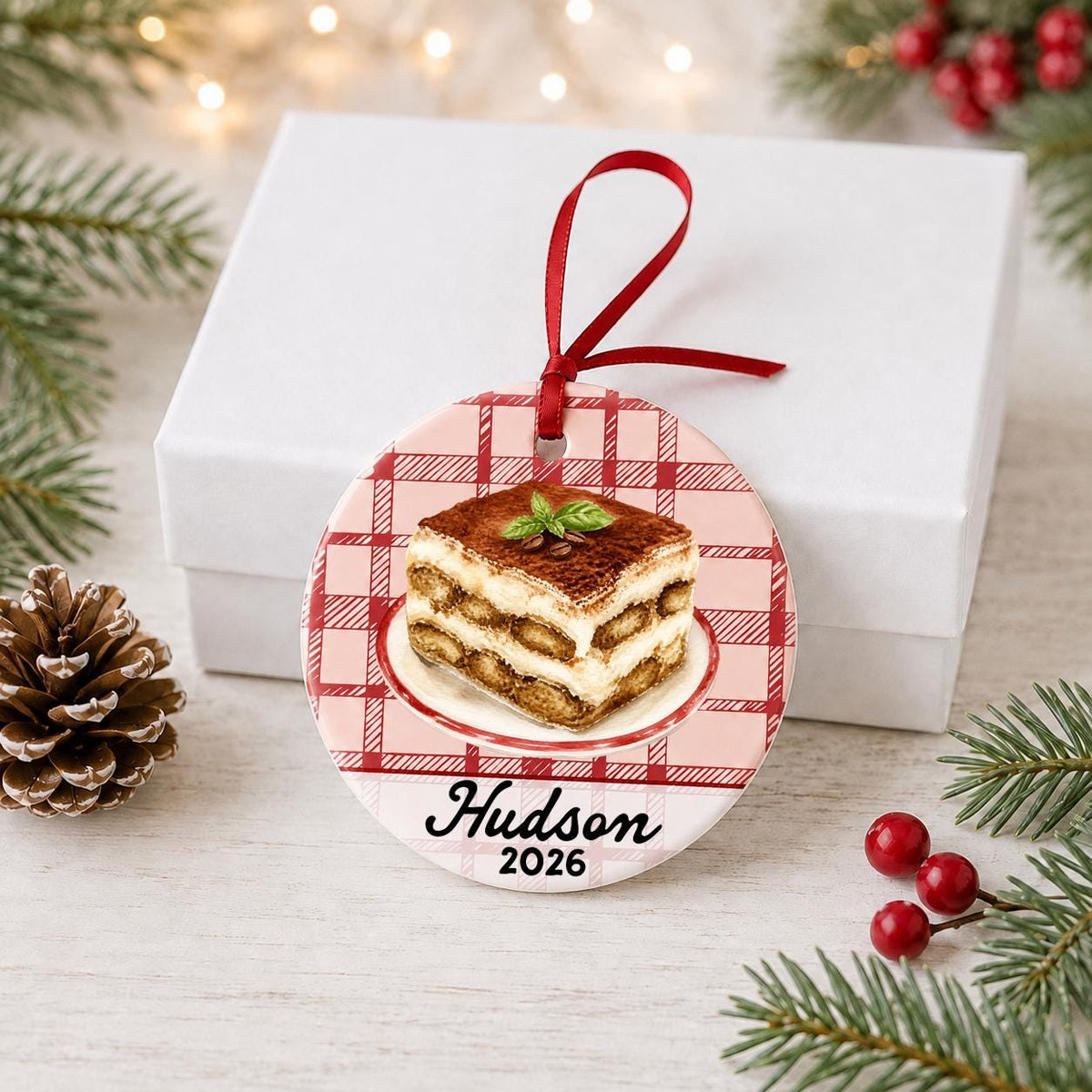 Personalized tiramisu ceramic ornament with name and year on red plaid in front of a white gift box with pinecones and red berries