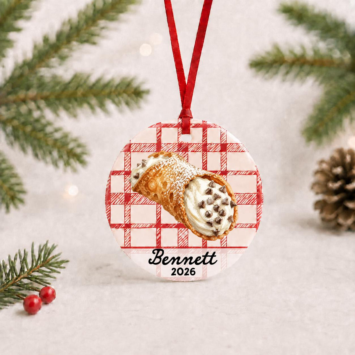 Personalized cannoli ceramic ornament with name and year on red plaid with red ribbon on a white surface with pine branches and pinecones