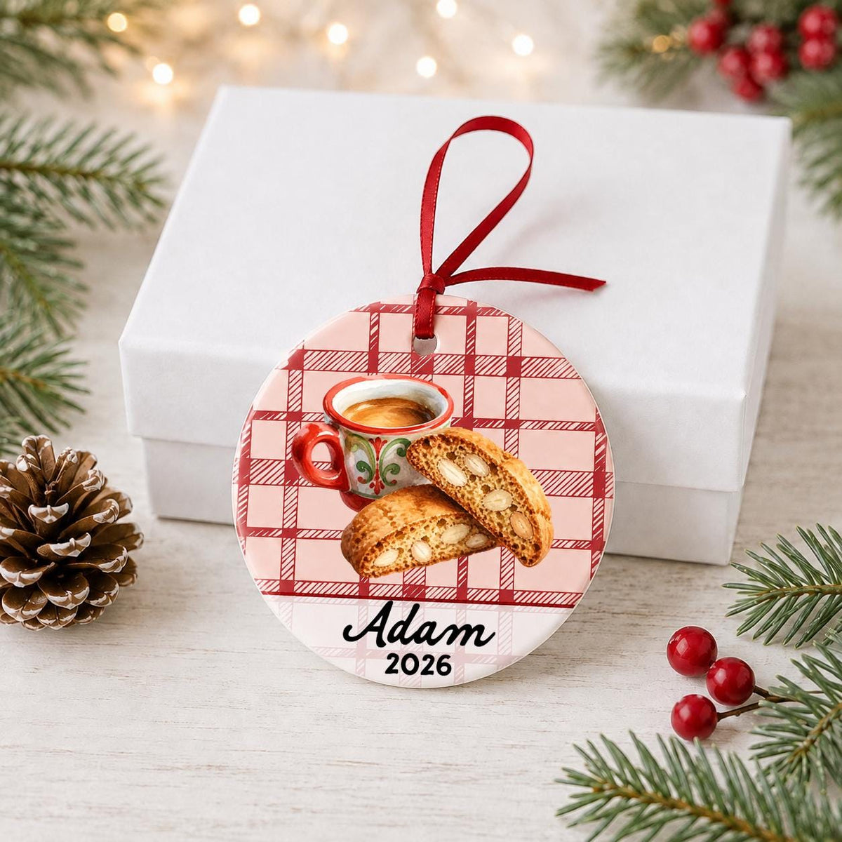 Personalized espresso and biscotti ceramic ornament with name and year on red plaid in front of a white gift box with pinecones and red berries