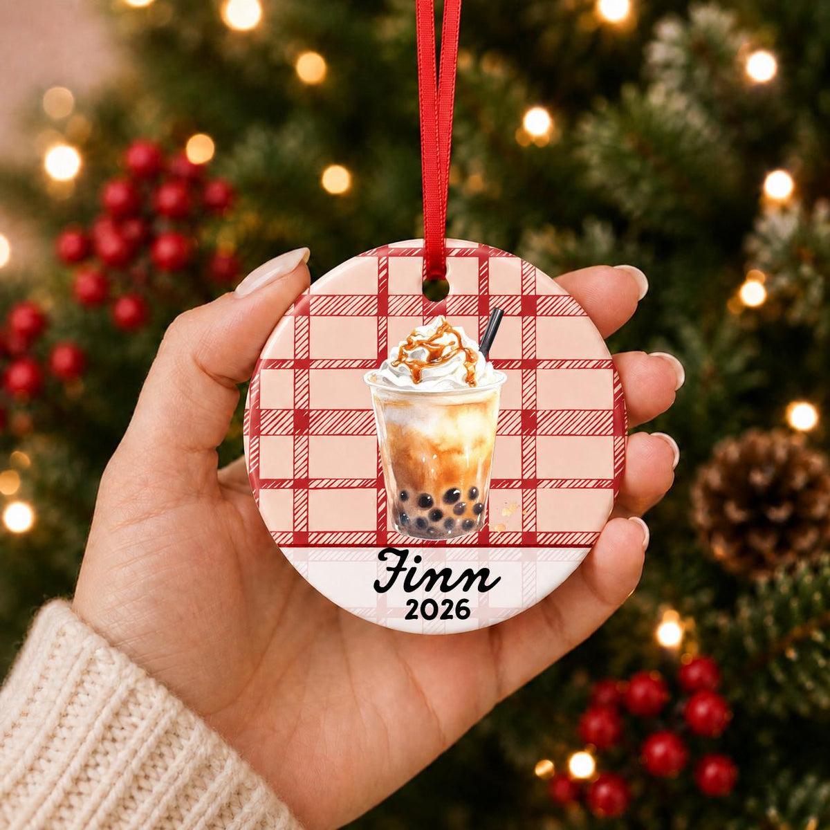 Hand holding the personalized brown sugar boba ceramic ornament with name and year on red plaid in front of a Christmas tree