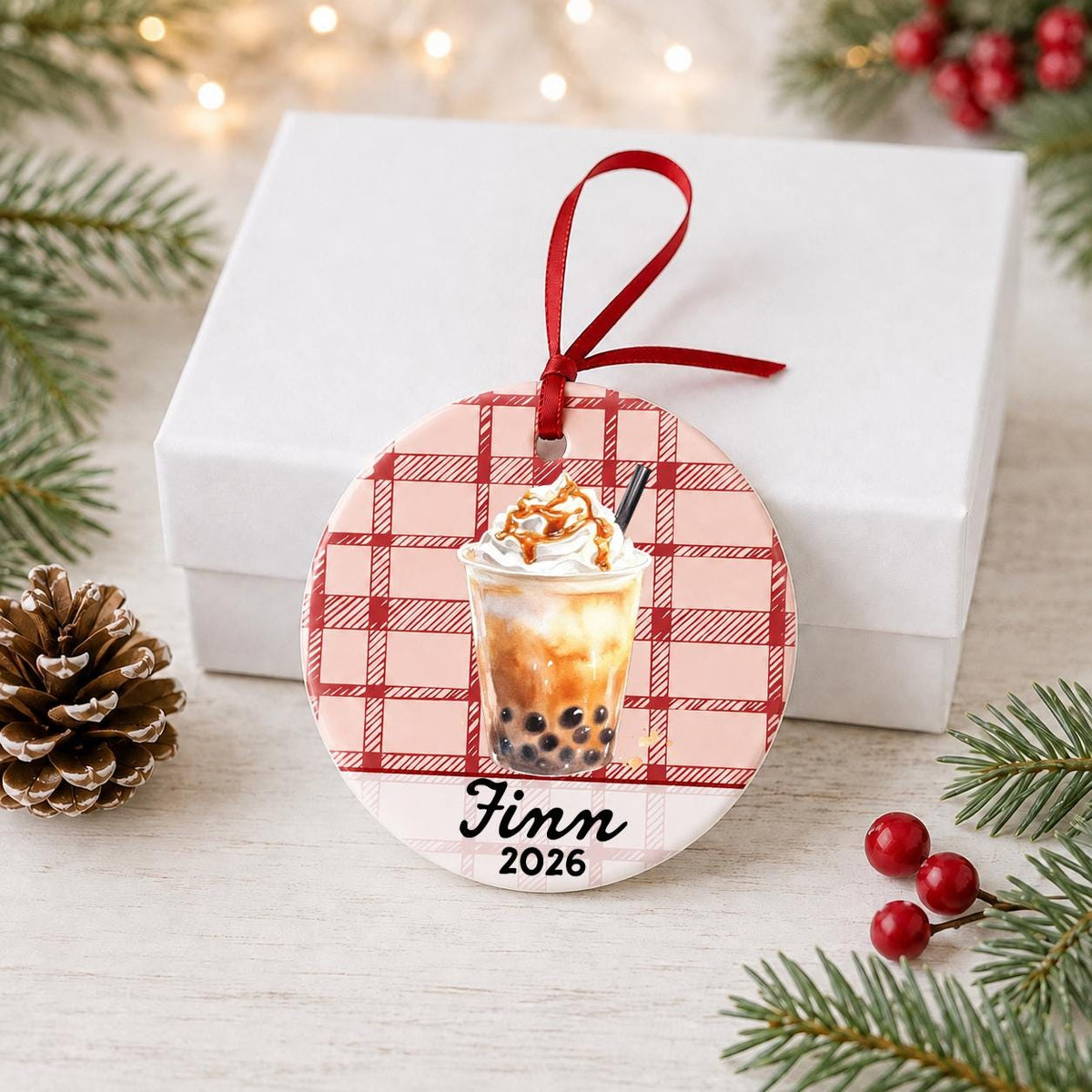 Personalized brown sugar boba ceramic ornament with name and year on red plaid in front of a white gift box with pinecones and red berries