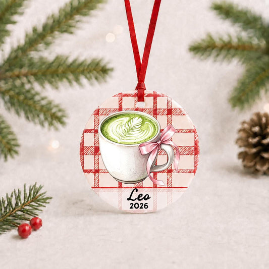 Personalized matcha latte ceramic ornament with name and year on red plaid with red ribbon on a white surface with pine branches and pinecones