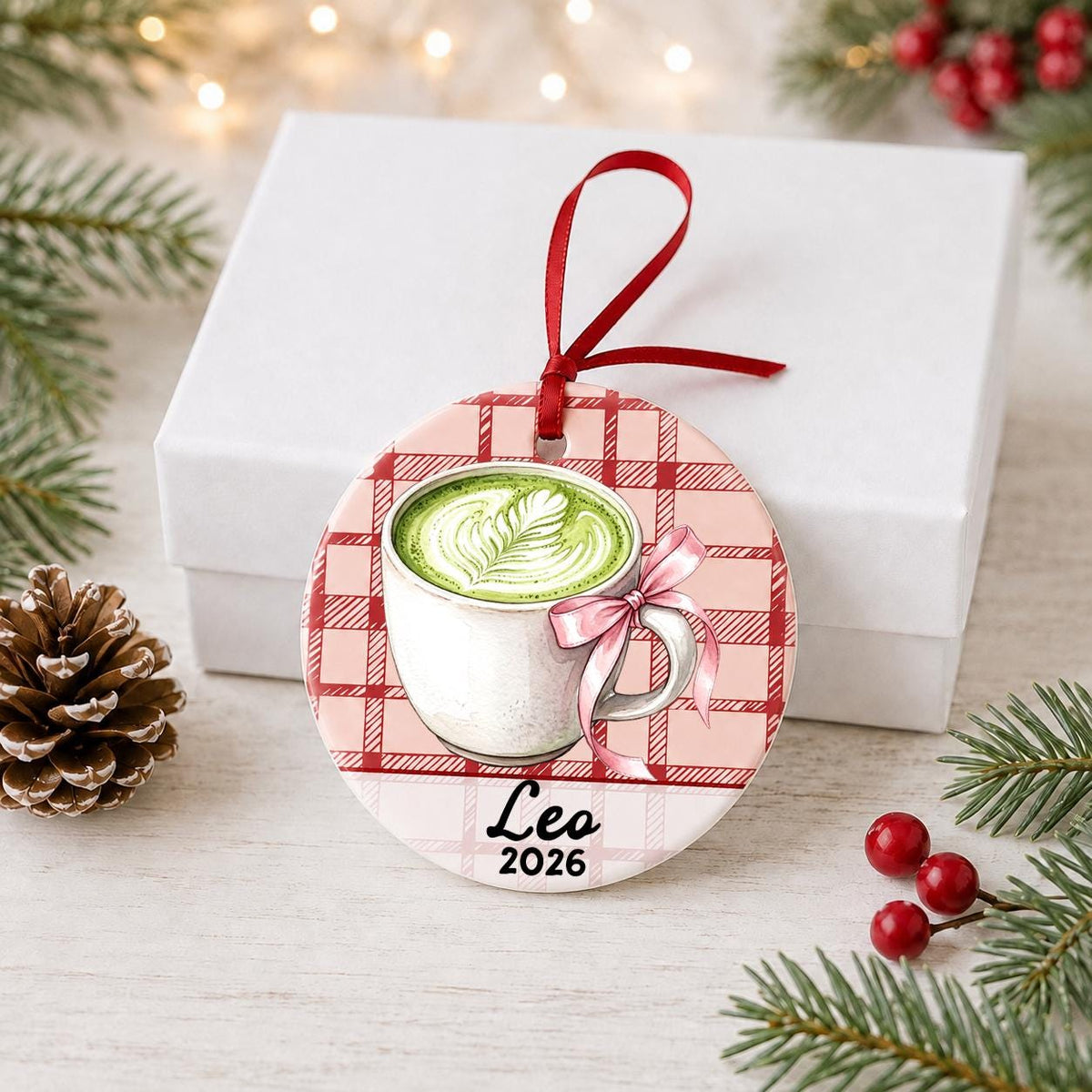 Personalized matcha latte ceramic ornament with name and year on red plaid in front of a white gift box with pinecones and red berries