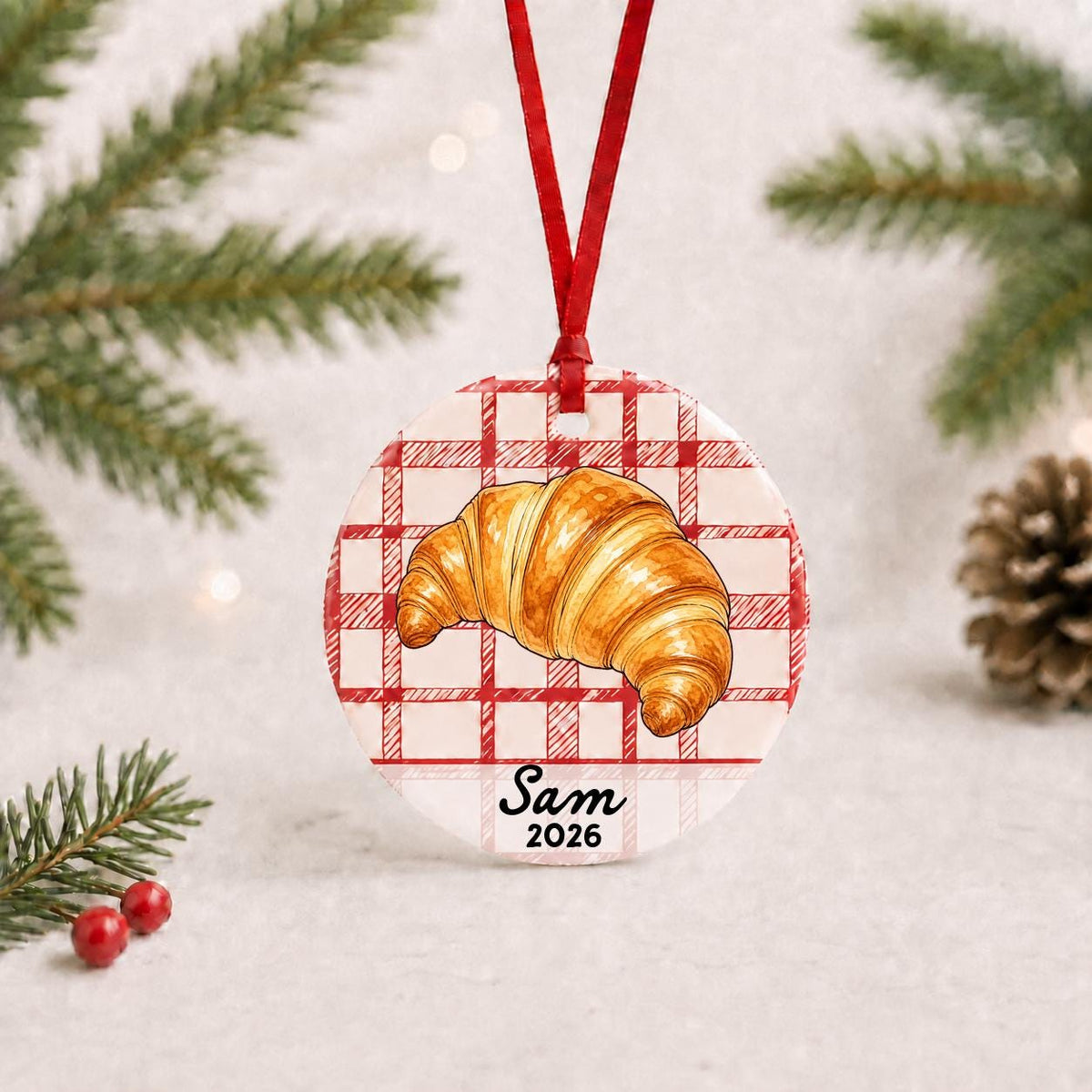 Personalized croissant ceramic ornament with name and year on red plaid with red ribbon on a white surface with pine branches and pinecones