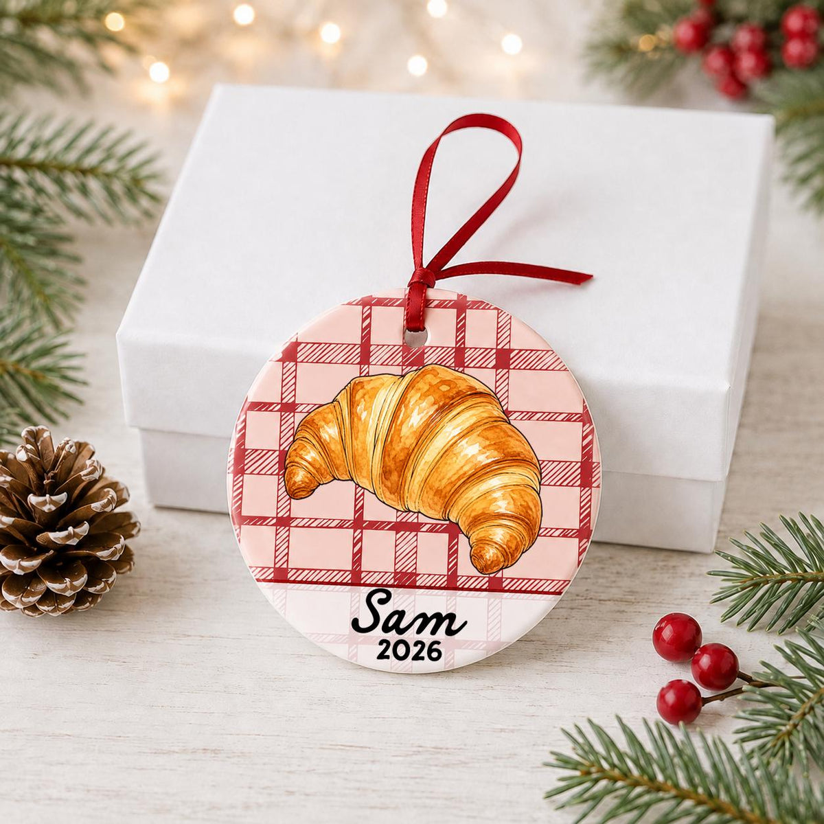 Personalized croissant ceramic ornament with name and year on red plaid in front of a white gift box with pinecones and red berries