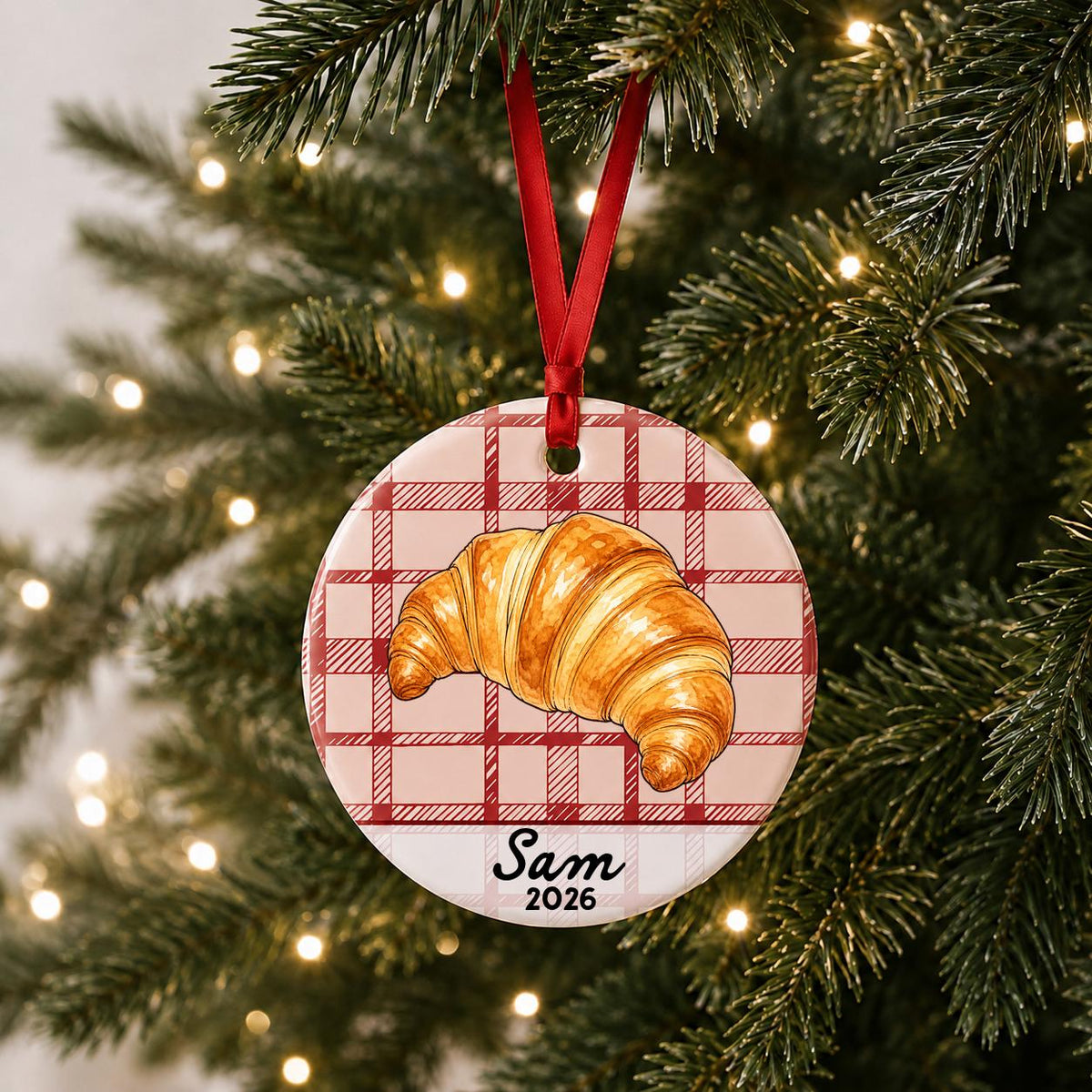 Personalized croissant ceramic ornament with name and year on red plaid hanging on a lit Christmas tree