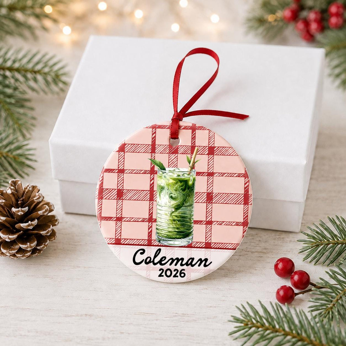Personalized iced matcha ceramic ornament with name and year on red plaid in front of a white gift box with pinecones and red berries