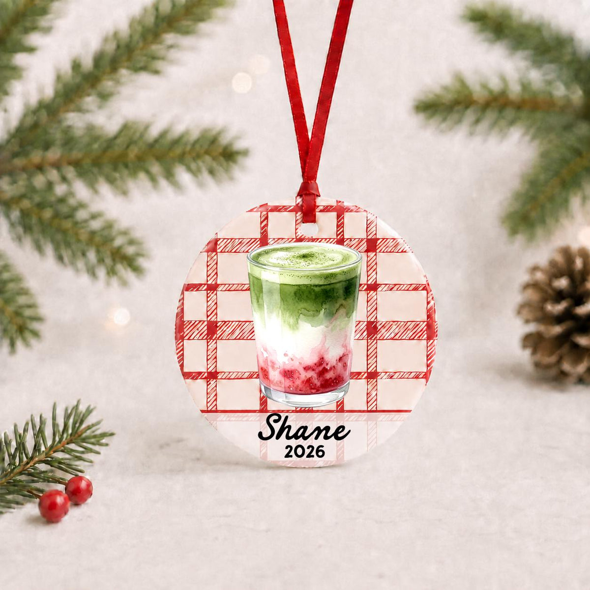 Personalized strawberry matcha latte ceramic ornament with name and year on red plaid with red ribbon on a white surface with pine branches and pinecones