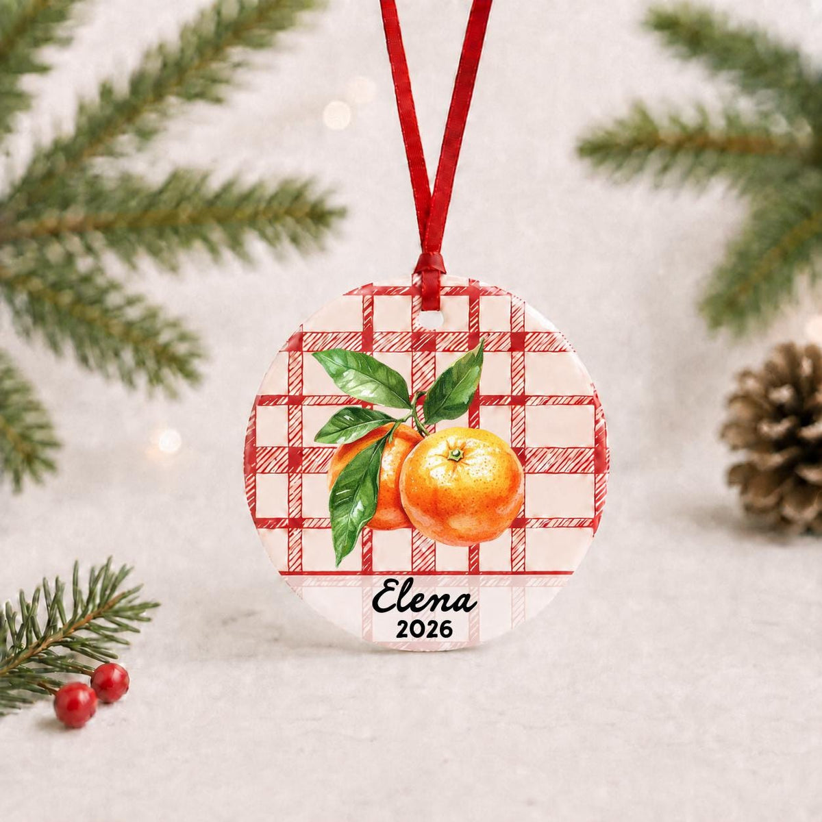 Personalized clementine ceramic ornament with name and year on red plaid with red ribbon on a white surface with pine branches and pinecones