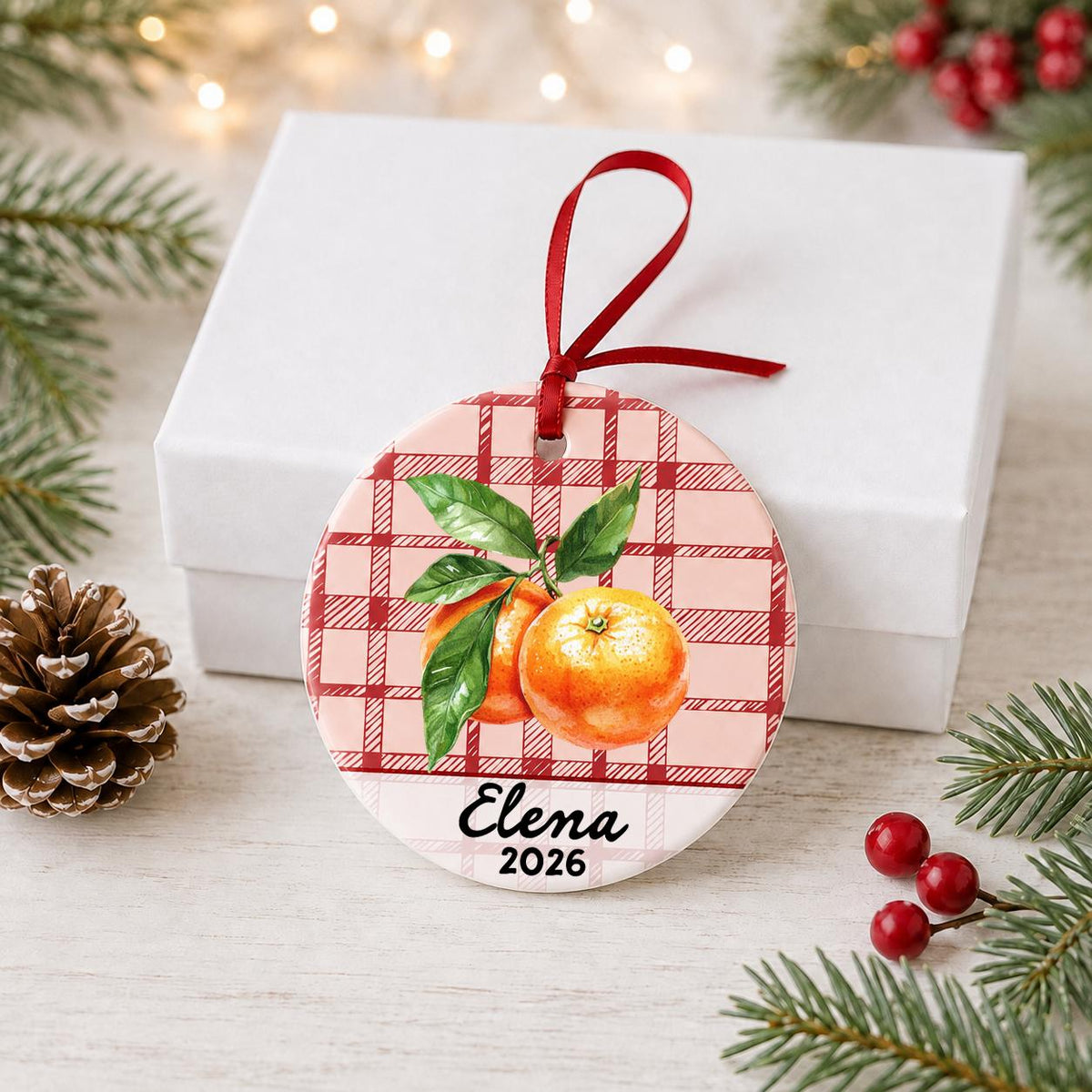 Personalized clementine ceramic ornament with name and year on red plaid in front of a white gift box with pinecones and red berries