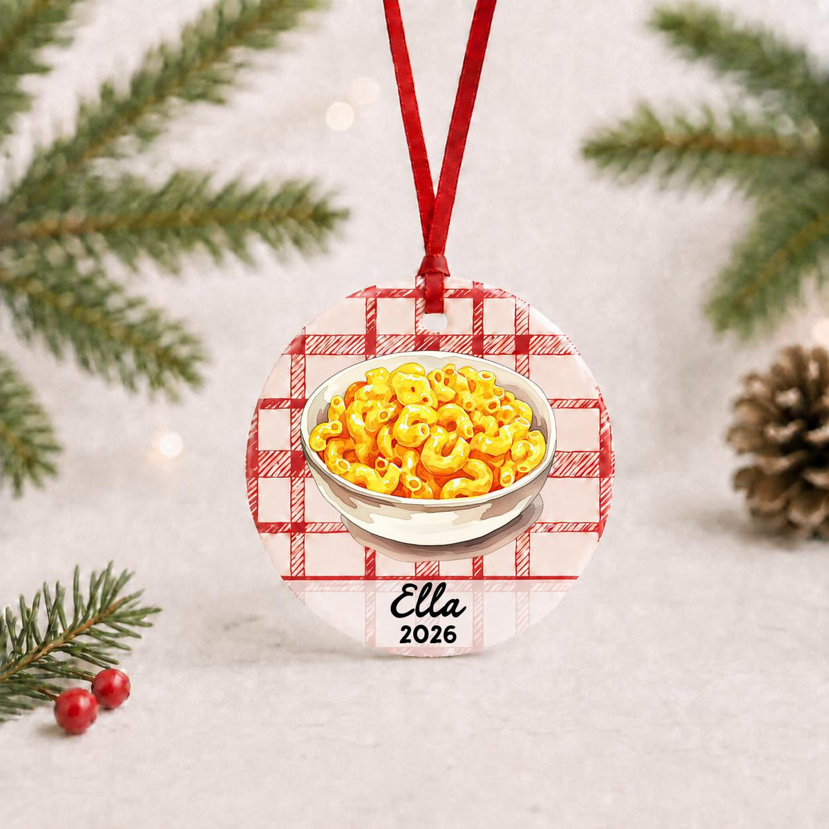 Personalized mac and cheese ceramic ornament with name and year on red plaid with red ribbon on a white surface with pine branches and pinecones