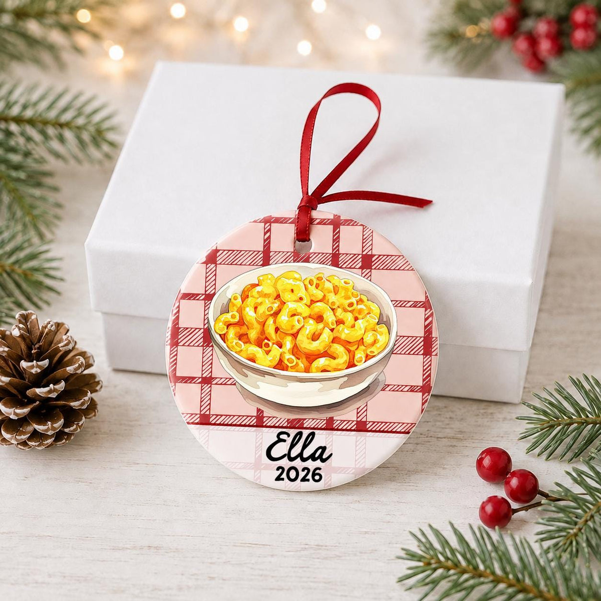 Personalized mac and cheese ceramic ornament with name and year on red plaid in front of a white gift box with pinecones and red berries