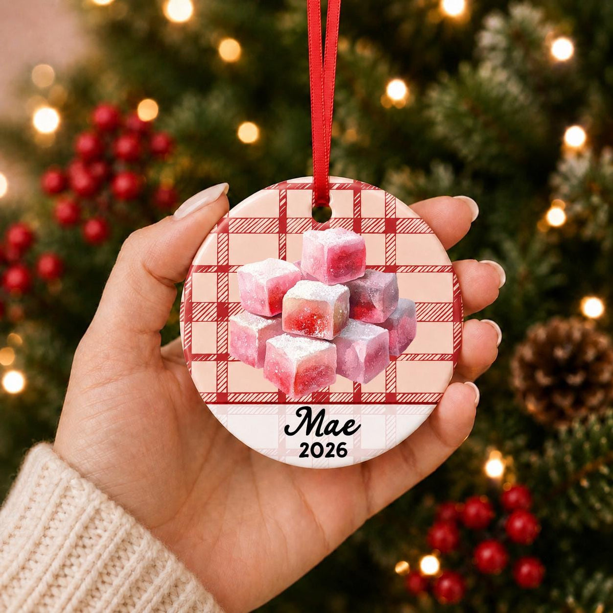 Hand holding the personalized turkish delight ceramic ornament with name and year on red plaid in front of a Christmas tree