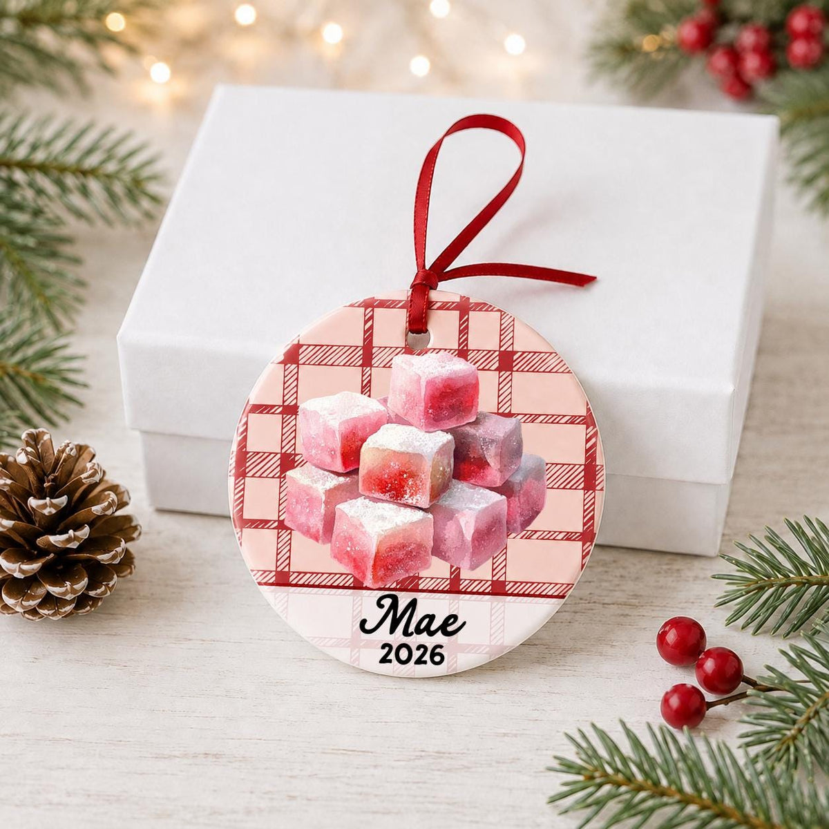 Personalized turkish delight ceramic ornament with name and year on red plaid in front of a white gift box with pinecones and red berries