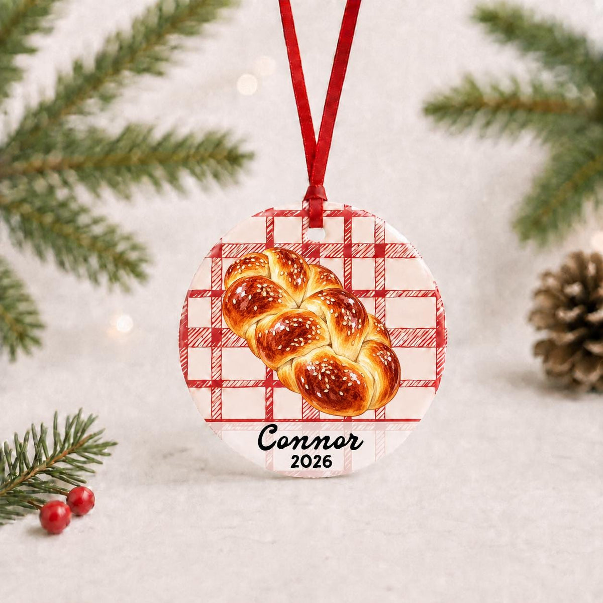 Personalized challah bread ceramic ornament with name and year on red plaid with red ribbon on a white surface with pine branches and pinecones