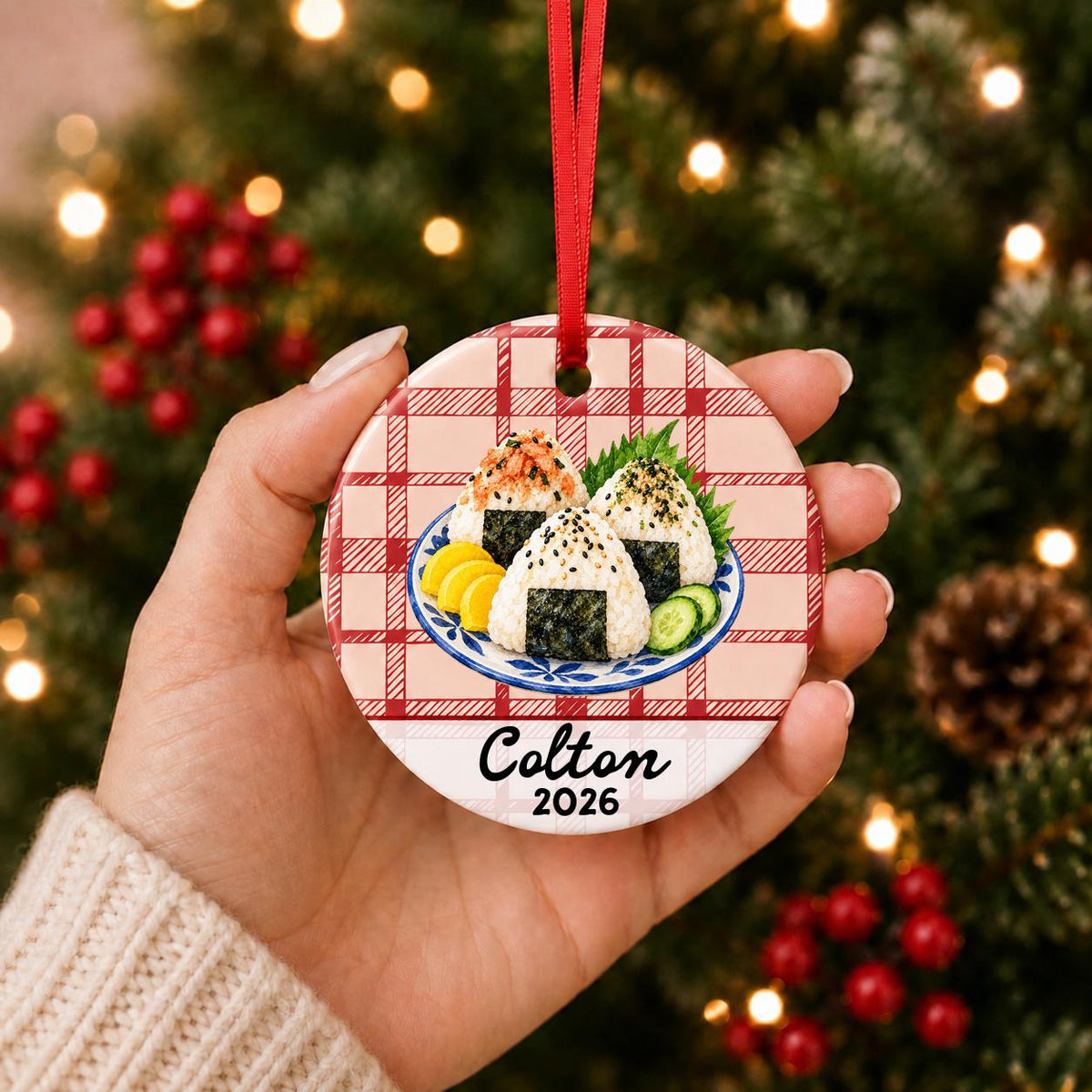 Hand holding the personalized onigiri ceramic ornament with name and year on red plaid in front of a Christmas tree