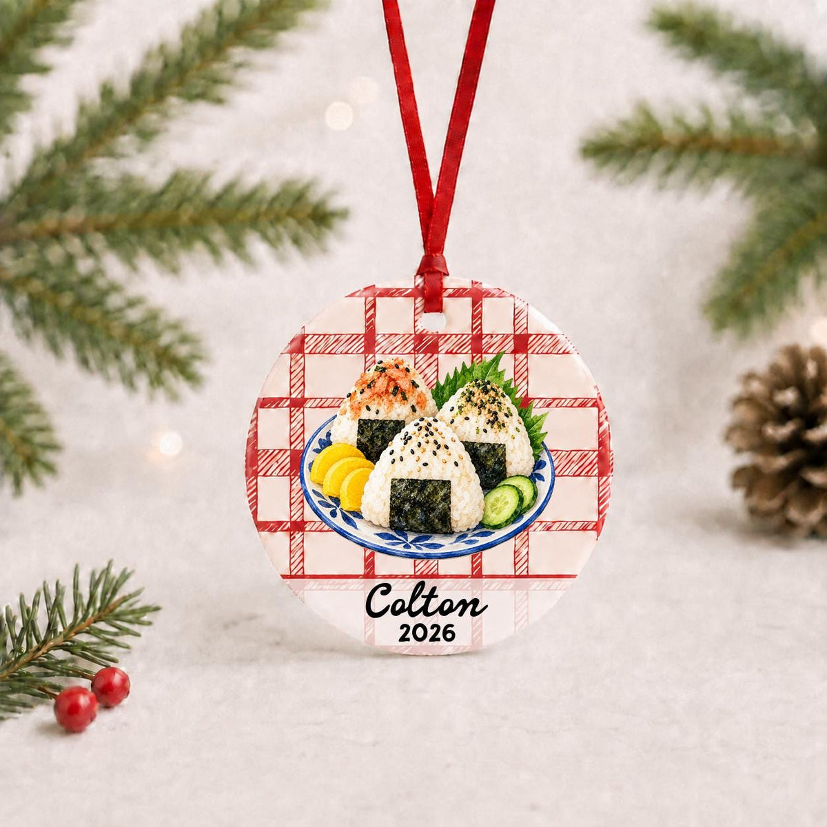 Personalized onigiri ceramic ornament with name and year on red plaid with red ribbon on a white surface with pine branches and pinecones
