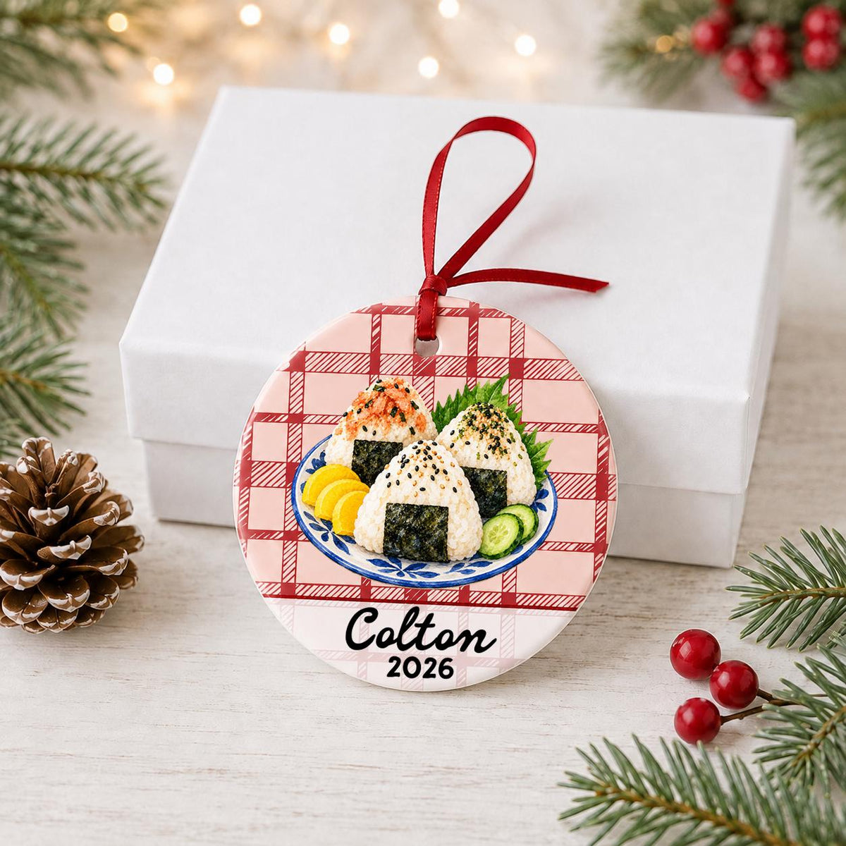Personalized onigiri ceramic ornament with name and year on red plaid in front of a white gift box with pinecones and red berries