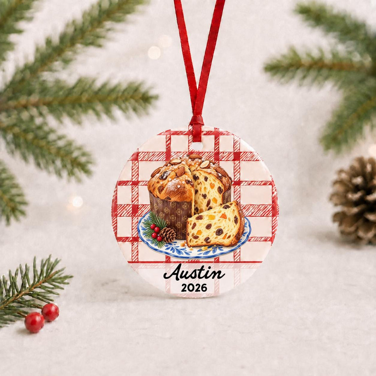 Personalized panettone ceramic ornament with name and year on red plaid with red ribbon on a white surface with pine branches and pinecones