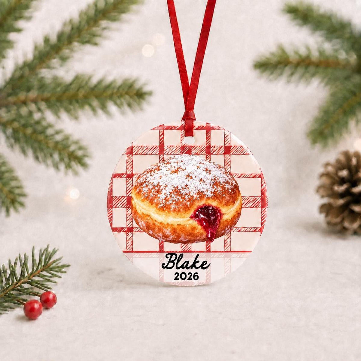 Personalized paczki ceramic ornament with name and year on red plaid with red ribbon on a white surface with pine branches and pinecones