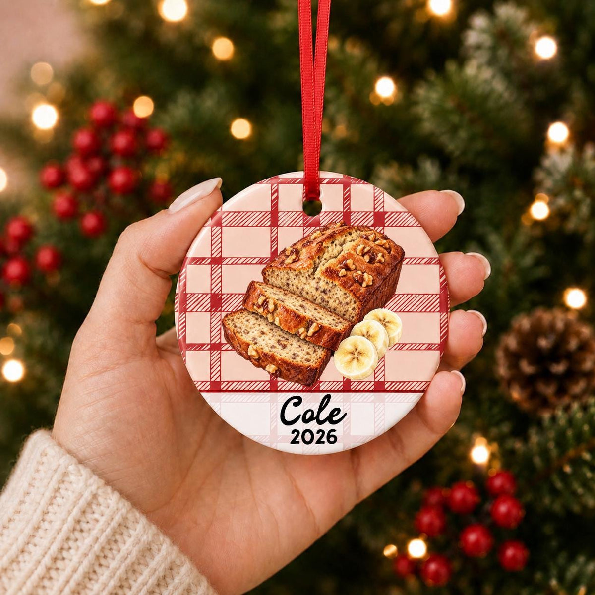 Hand holding the personalized banana bread ceramic ornament with name and year on red plaid in front of a Christmas tree