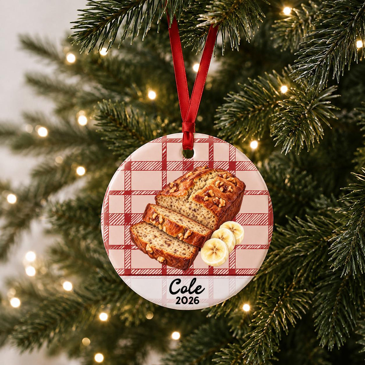 Personalized banana bread ceramic ornament with name and year on red plaid hanging on a lit Christmas tree