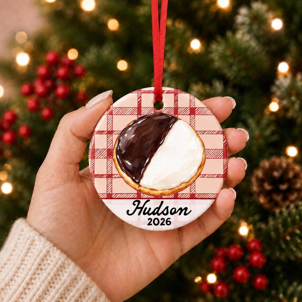 Hand holding the personalized black and white cookie ceramic ornament with name and year on red plaid in front of a Christmas tree