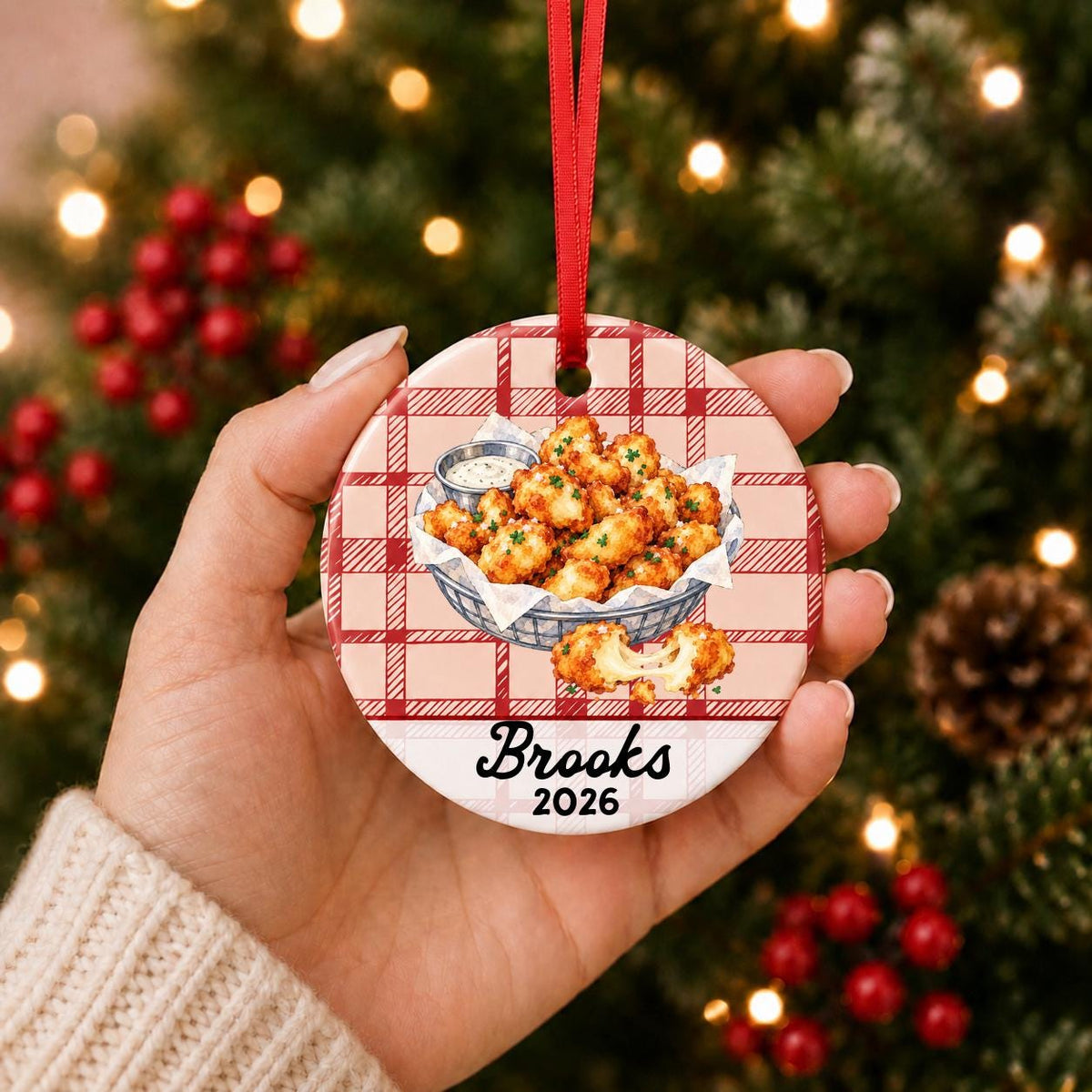 Hand holding the personalized cheese curds ceramic ornament with name and year on red plaid in front of a Christmas tree