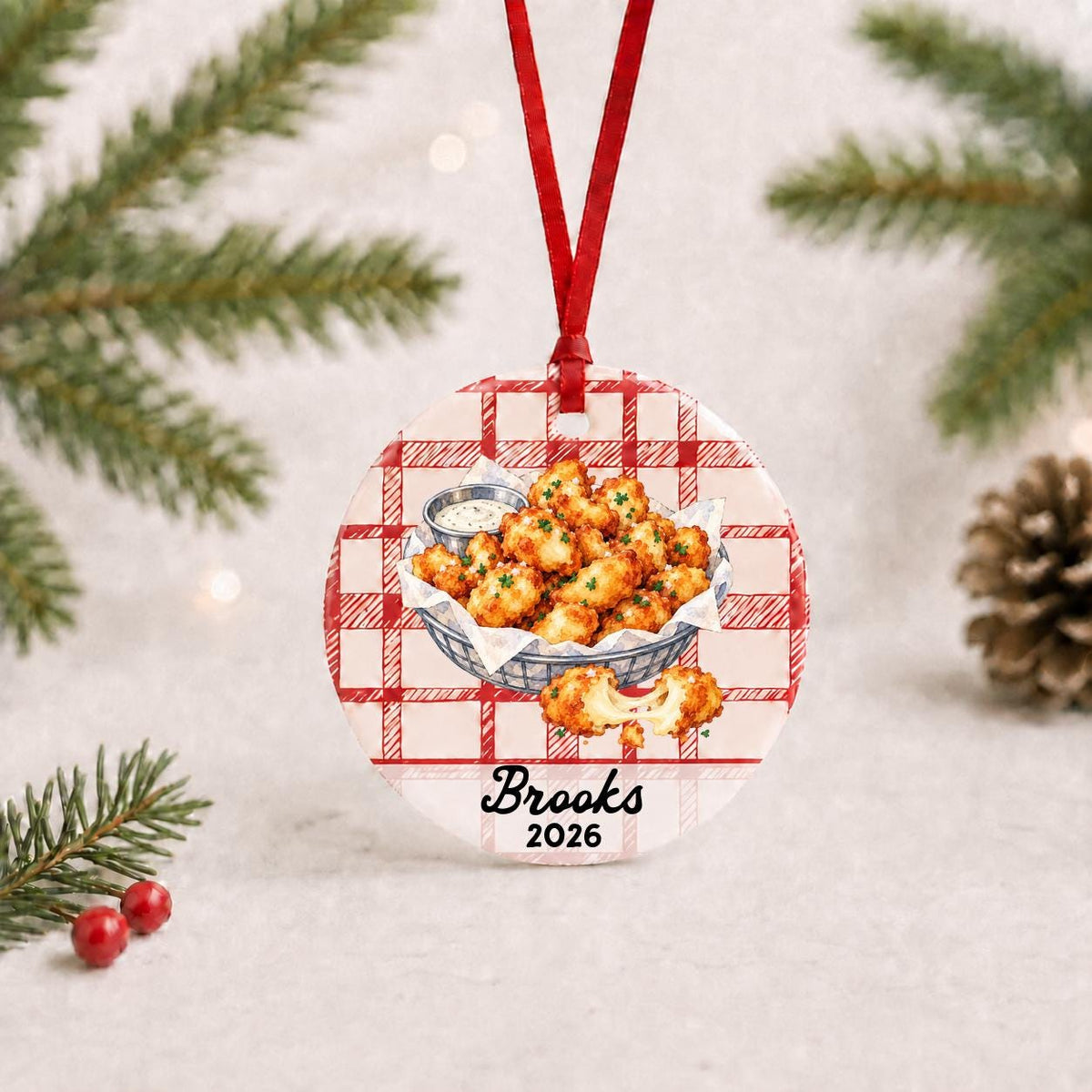 Personalized cheese curds ceramic ornament with name and year on red plaid with red ribbon on a white surface with pine branches and pinecones