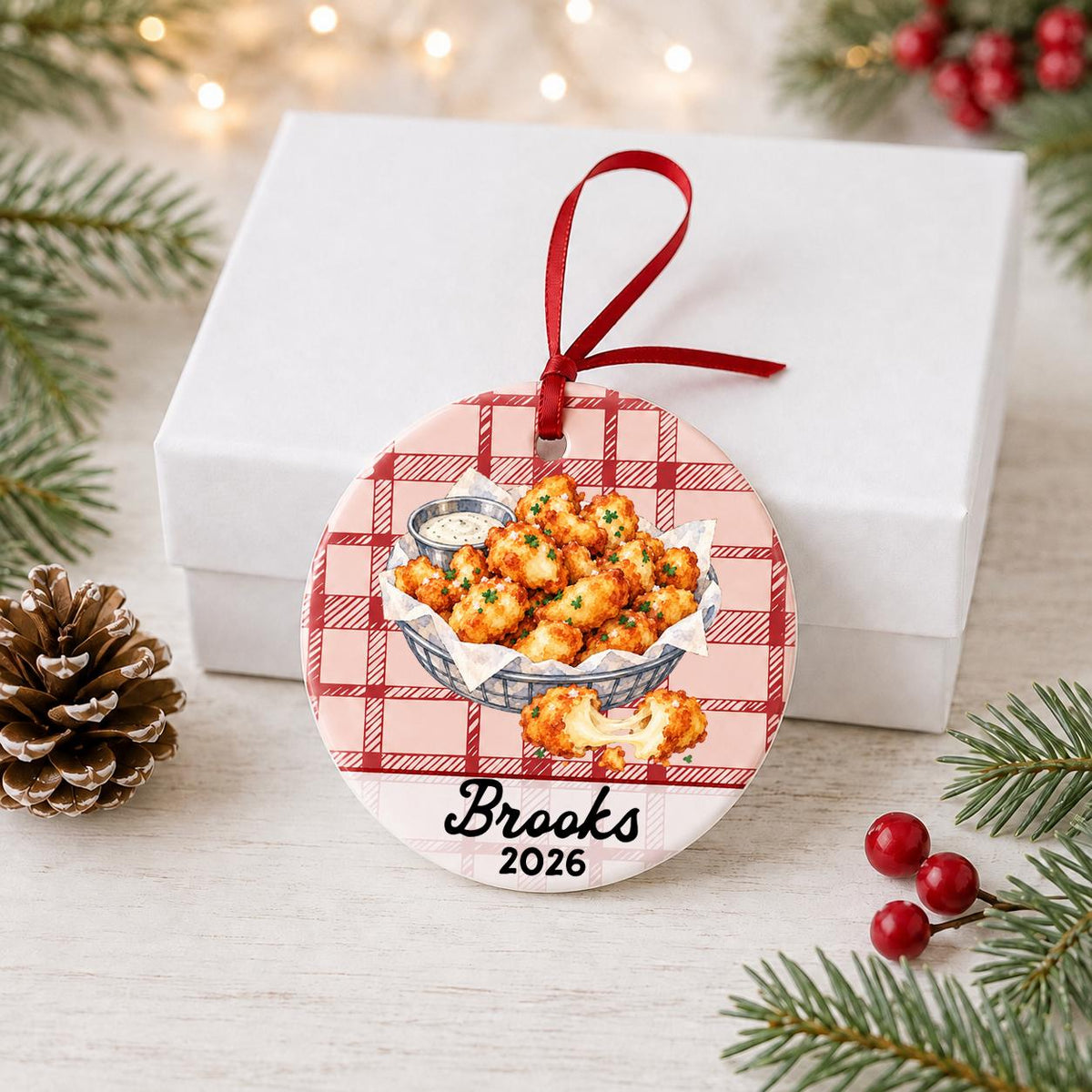 Personalized cheese curds ceramic ornament with name and year on red plaid in front of a white gift box with pinecones and red berries