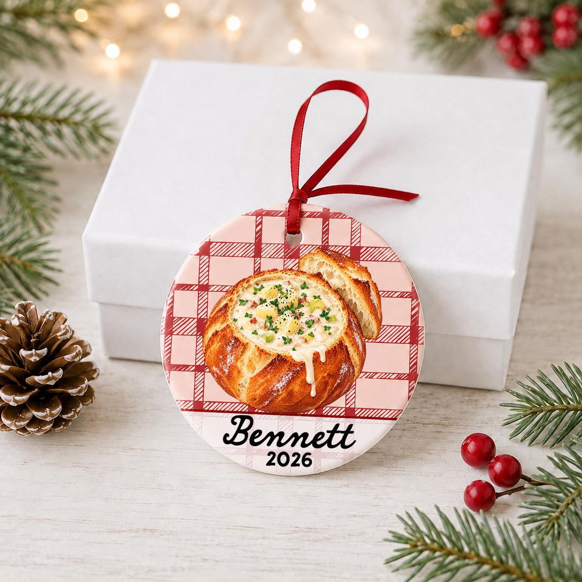 Personalized clam chowder bread bowl ceramic ornament with name and year on red plaid in front of a white gift box with pinecones and red berries