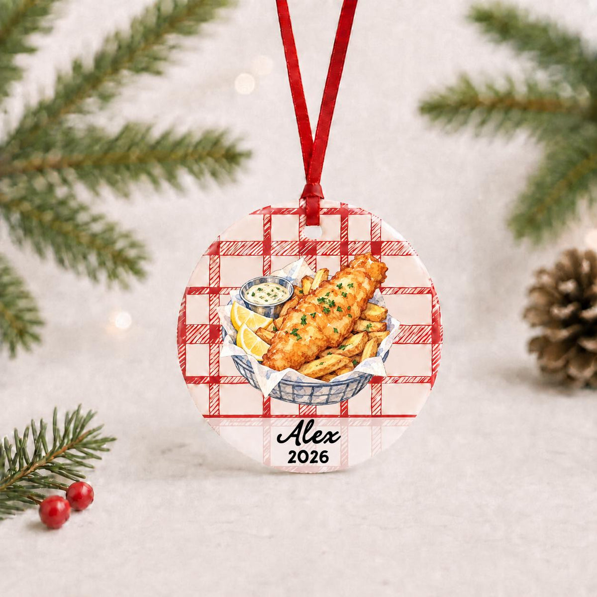 Personalized fish and chips ceramic ornament with name and year on red plaid with red ribbon on a white surface with pine branches and pinecones