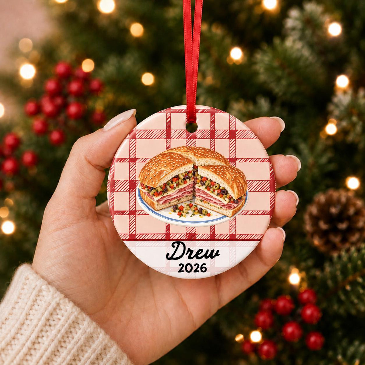 Hand holding the personalized muffuletta ceramic ornament with name and year on red plaid in front of a Christmas tree