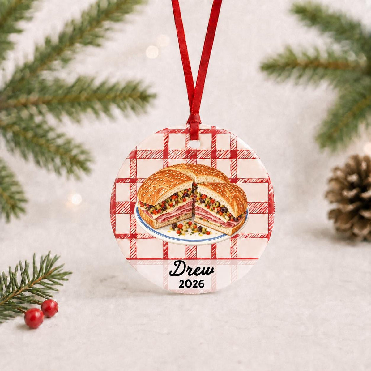 Personalized muffuletta ceramic ornament with name and year on red plaid with red ribbon on a white surface with pine branches and pinecones
