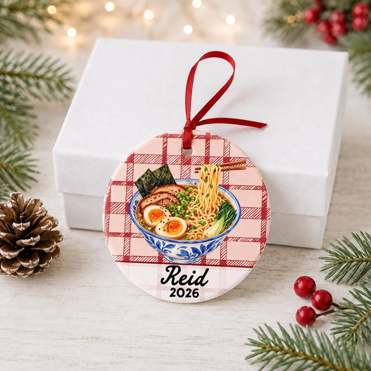 Personalized ramen ceramic ornament with name and year on red plaid in front of a white gift box with pinecones and red berries