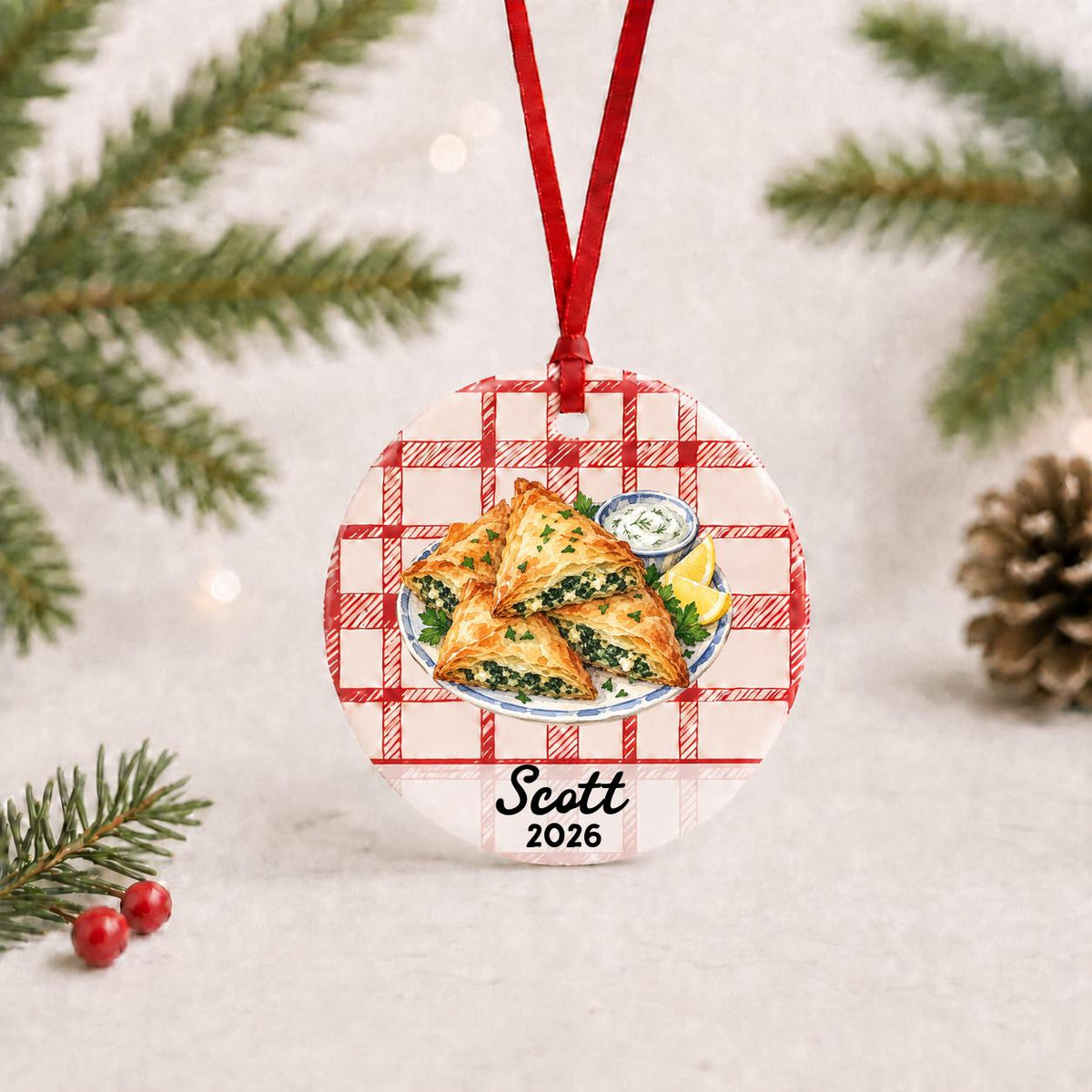 Personalized spanakopita ceramic ornament with name and year on red plaid with red ribbon on a white surface with pine branches and pinecones