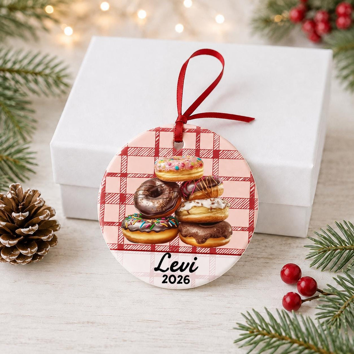 Personalized donuts ceramic ornament with name and year on red plaid in front of a white gift box with pinecones and red berries
