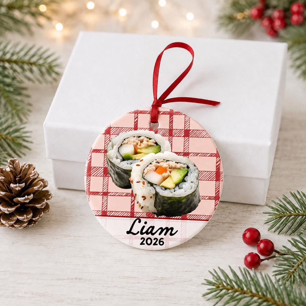 Personalized sushi ceramic ornament with name and year on red plaid in front of a white gift box with pinecones and red berries