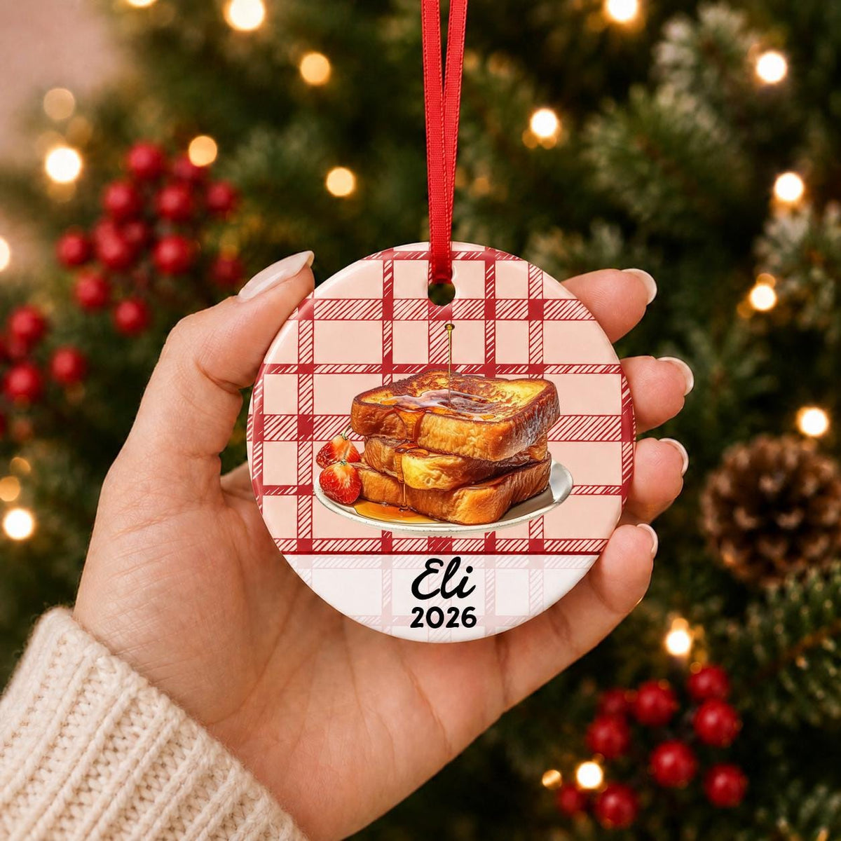 Hand holding the personalized French toast ceramic ornament with name and year on red plaid in front of a Christmas tree