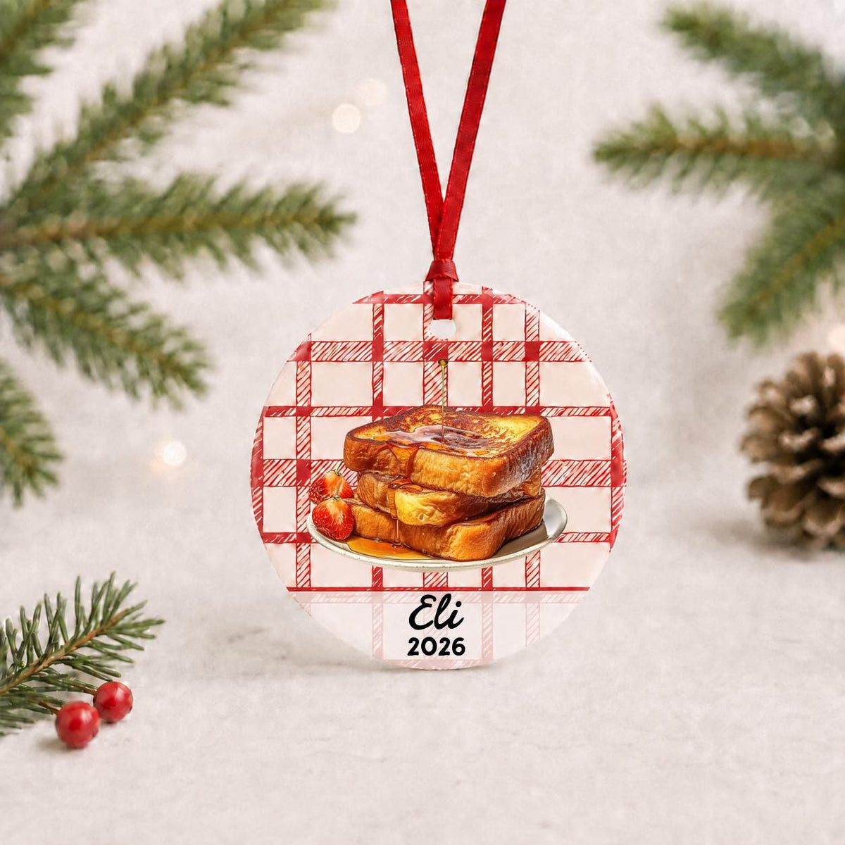 Personalized French toast ceramic ornament with name and year on red plaid with red ribbon on a white surface with pine branches and pinecones
