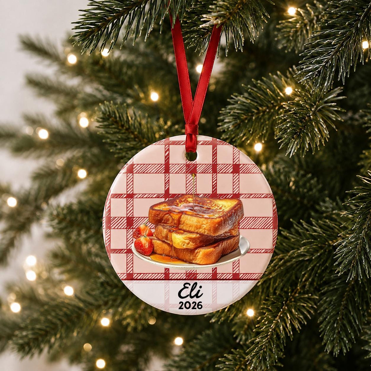 Personalized French toast ceramic ornament with name and year on red plaid hanging on a lit Christmas tree