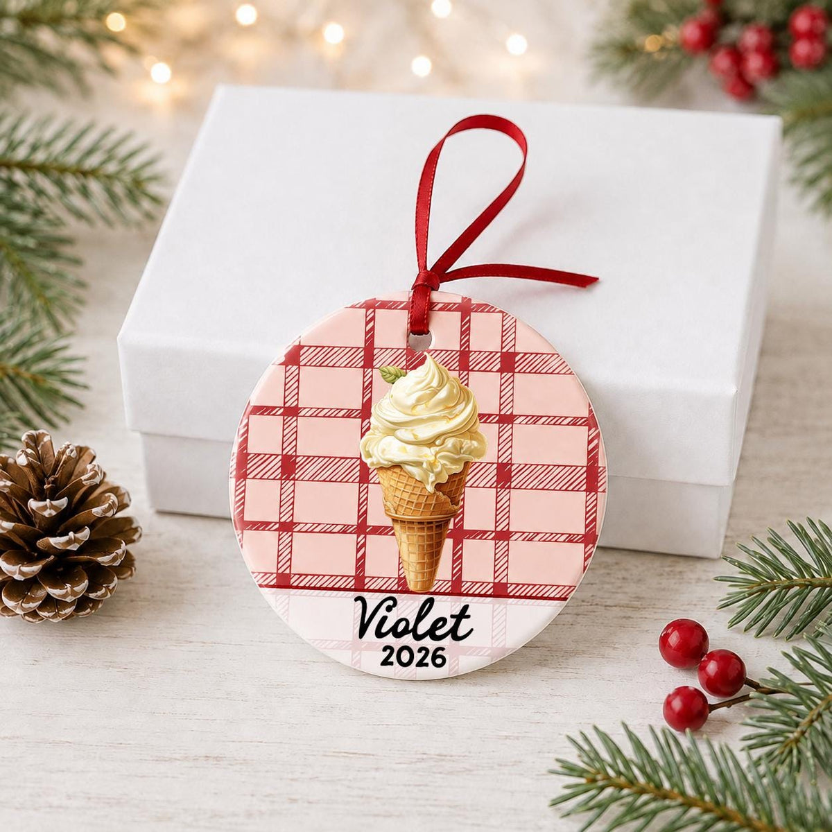Personalized soft serve ice cream ceramic ornament with name and year on red plaid in front of a white gift box with pinecones and red berries