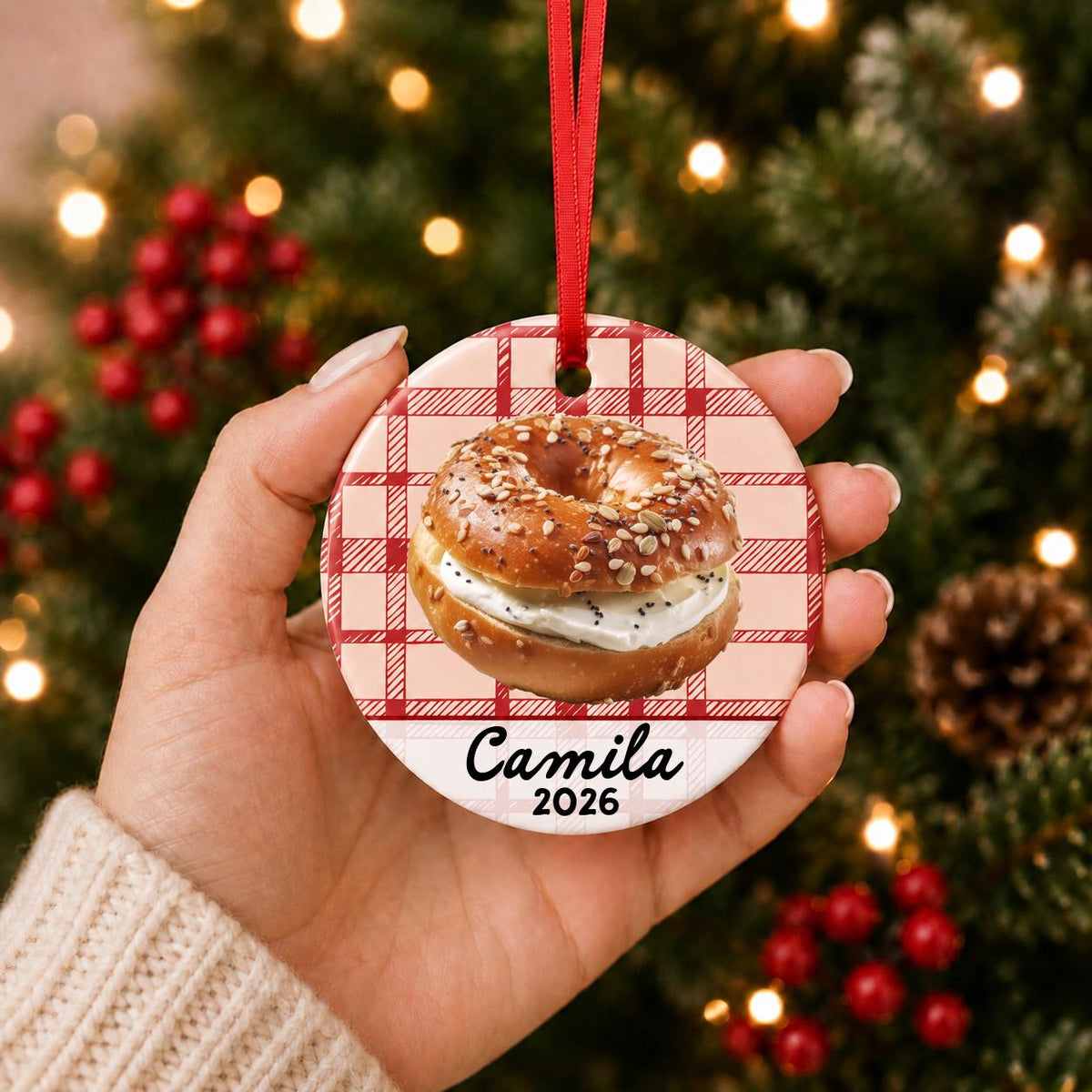 Hand holding the personalized bagel ceramic ornament with name and year on red plaid in front of a Christmas tree
