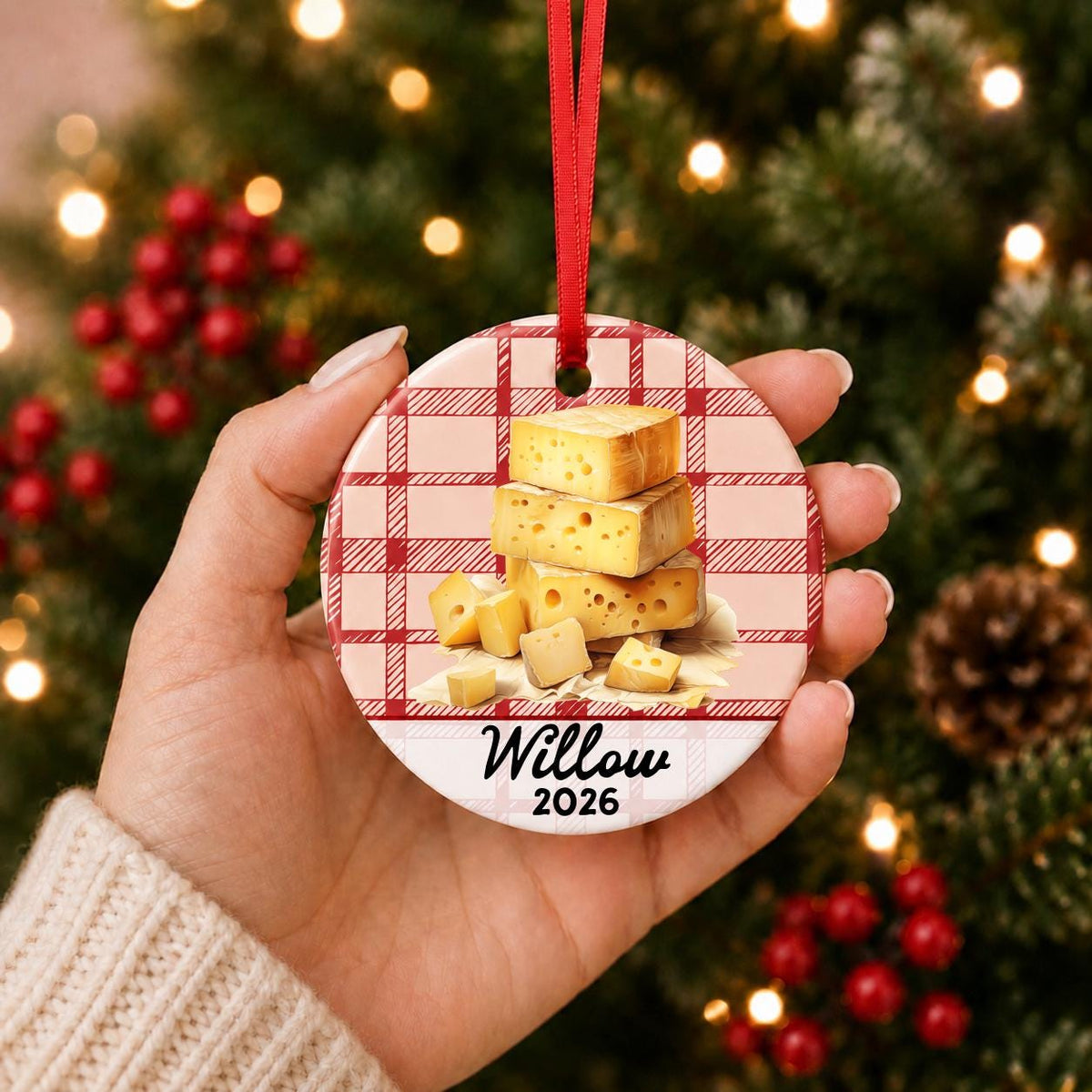 Hand holding the personalized Swiss cheese ceramic ornament with name and year on red plaid in front of a Christmas tree