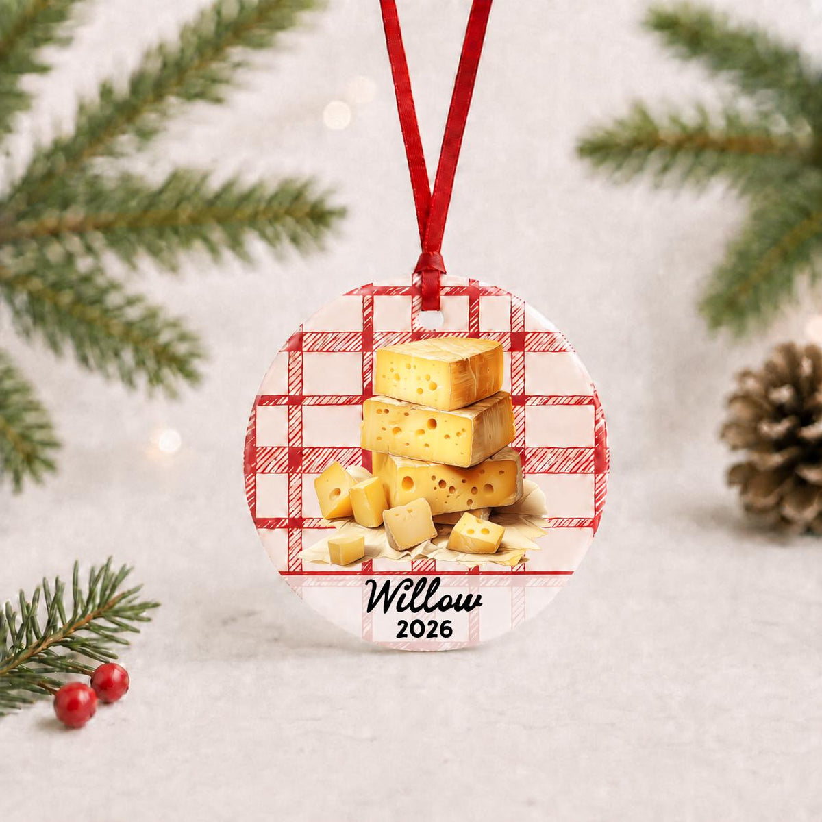 Personalized Swiss cheese ceramic ornament with name and year on red plaid with red ribbon on a white surface with pine branches and pinecones