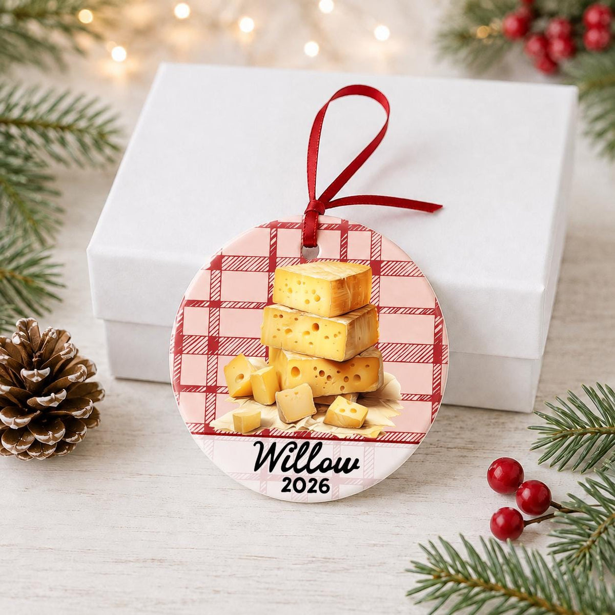 Personalized Swiss cheese ceramic ornament with name and year on red plaid in front of a white gift box with pinecones and red berries