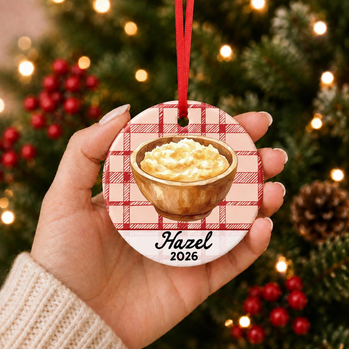 Hand holding the personalized mashed potatoes ceramic ornament with name and year on red plaid in front of a Christmas tree